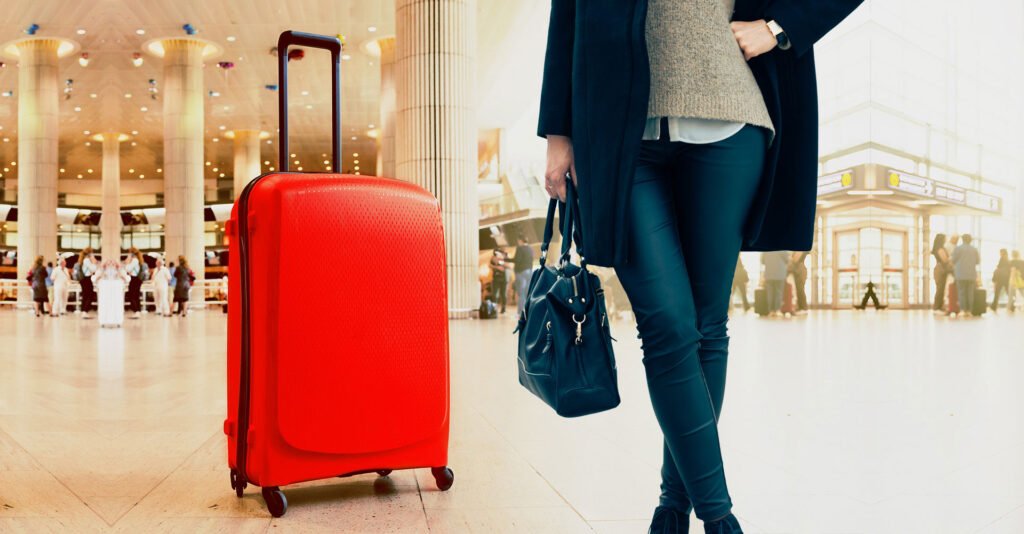 A person in stylish winter attire holding a dark blue handbag, standing beside a bright red rolling suitcase in an airport terminal.