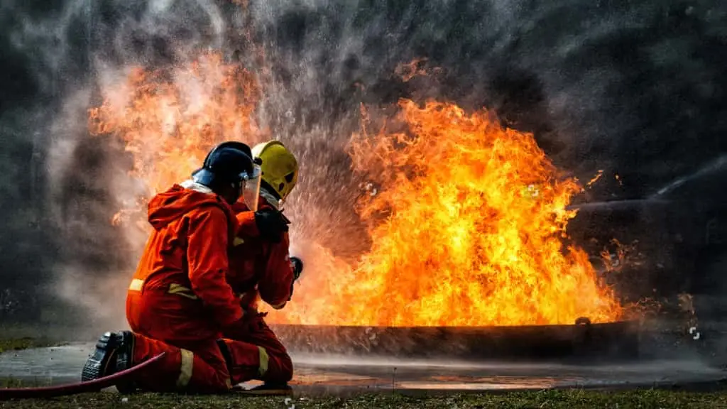 Two firefighters in red uniforms and helmets extinguishing a large fire with a hose.